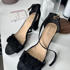 Svarta klackar och volang - Stilrena svarta sandaler med bred klack och öppen tå. Skorna har en snygg volangdetalj framtill och justerbart spänne vid ankeln i guld. Materialet är mockaliknande 🌸