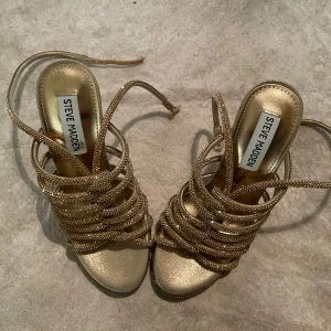 Steve Madden guldiga sandaler med klack - Glittriga guldiga sandaler från Steve Madden med höga klackar och tunna remmar täckta av små stenar. Öppen tå och justerbar rem runt vristen. Ovandel i tyg och glas, foder i syntet. Perfekta för dig som vill sticka ut.