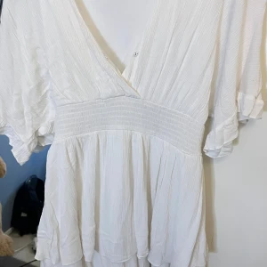 Vit omlottklänning med volang - Superfin vit kortklänning med omlott och v-ringning. Klänningen har bred resår i midjan, volangdetaljer nertill och korta, lösa ärmar med lätt rynk. Perfekt för sommarens alla tillfällen och ger en luftig och fräsch känsla.