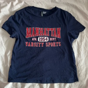 Mörkblå Manhattan t-shirt med tryck - Mörkblå t-shirt med rött och vitt tryck på bröstet där det står 'MANHATTAN 1954 VARSITY SPORTS'. Klassisk rund halsringning och korta ärmar. Skön och mjuk bomullskvalitet, perfekt för en sportig och avslappnad look.