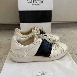 Valentino Garavani Rockstud sneakers - Snygga vita Valentino Garavani Rockstud sneakers med svart band och ikoniska nitar. Skorna har rund tå, snörning och är tillverkade i skinn. Kan tänka mig att gå ner i pris så tar emot prisförslag! Box och dustbag ingår💗