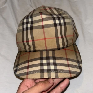 Burberry  keps i beige och svart - Snygg burberry keps med klassiskt rutigt mönster i beige, svart, vitt och röda linjer. Justerbar rem baktill och tydlig Burberry London England-patch.  Den. Använt 5 gånger max 