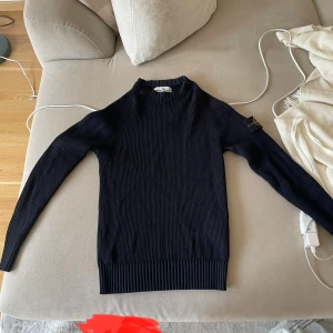 Mörkblå stickad tröja Stone Island - Säljer en mörkblå ribbstickad tröja från Stone Island med klassisk patch på ärmen. Tröjan har lång ärm, högre krage och är i ett mjukt, stretchigt material som sitter snyggt på kroppen. Perfekt för dig som gillar stilren och sportig design.