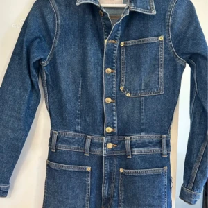 Jeans jumpsuit från Zara 70s Work Wear - Snygg jeans jumpsuit från Zara 70s Work Wear Capsule. Klassisk blå denim med raka ben, stora fickor fram och bak, bälte i midjan och guldiga knappar. Långärmad och med krage för en cool vintagekänsla. Perfekt för dig som gillar retrostil eller bara vill ha en fin jumpsuit.🥰