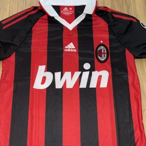 AC Milan Beckham 32 Adidas fotbollströja - Säljer en klassisk AC Milan fotbollströja från Adidas med Beckham och nummer 32 på ryggen. Tröjan är röd och svart med vertikala ränder, vit krage och klubbmärke på bröstet. Materialet är lätt och andas, perfekt för match eller träning.