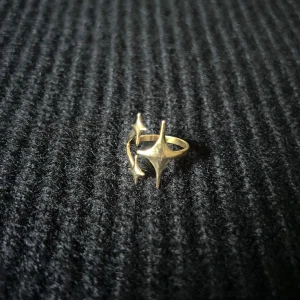 Halo Nocturne Doré – Starfall Embrace Ring - Justerbar ring i rostfritt stål med guldplätering, rost- och nickelfri. Tre stjärnor i olika nivåer följer en mjuk båge runt fingret – en stilren design med känsla av rörelse och ljus. 