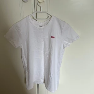 Vit t-shirt från Levi's - Klassisk vit t-shirt från Levi's med liten röd logotyp på bröstet. Enkel och stilren design med rund hals och korta ärmar. Perfekt basplagg i mjuk bomull som funkar till allt. Snygg att matcha med jeans eller shorts.