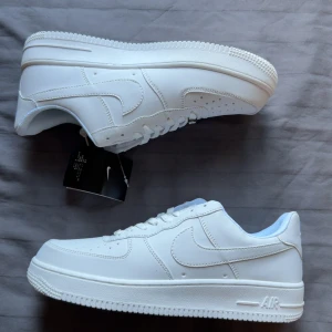 Nike Air Force 1 helvita sneakers - Klassiska Nike Air Force 1 sneakers i helvitt skinn med låg profil och rund tå. Skorna har vit sula, perforeringar på tån för ventilation och ikonisk Swoosh-logga på sidorna. Perfekta för dig som gillar en clean och tidlös look.