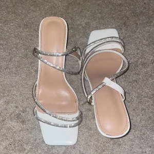 Vita sandaler med strass och klack - Snygga vita sandaler med smala remmar täckta av glittrande strass. Skorna har fyrkantig tå, öppen design och smal hög klack. Perfekta för dig som vill sticka ut med en elegant och trendig look. Materialet är syntet och de har en stilren, modern vibe.