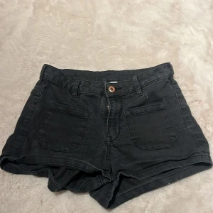 Svarta jeansshorts med hög midja - Snygga svarta jeansshorts med hög midja och klassisk femficksdesign. Shortsen har framfickor, bälteshällor och stängs med dragkedja och knapp. Perfekta till sommaren och lätta att matcha med allt.