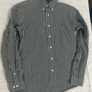 Randig skjorta Polo Ralph Lauren M - Snygg svart och vit randig skjorta från Polo Ralph Lauren i custom fit. Skjortan har klassisk krage, långa ärmar och knäppning framtill. Broderad lila logga på bröstet. Tillverkad i 100% bomull för en skön känsla.