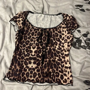 Leopardmönstrad blus med rosett - Snygg blus med leopardmönster i brunt och beige. Modellen har korta ärmar, svart spetskant vid urringningen och en söt rosett framtill. Tunt, flowy material som känns lätt och luftigt. Perfekt för dig som vill sticka ut med ett djurmönster.