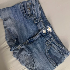 Blå jeansshorts med råa kanter - Snygga blå jeansshorts från H&M med råa, fransiga kanter och coola fickdetaljer bak med knappar. Klassisk femficksmodell och bälteshällor. Perfekta för varma dagar när du vill ha en avslappnad och trendig look.