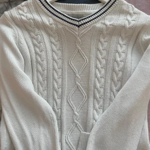 Vit stickad tröja från Cappuccini - Vit stickad tröja från Cappuccini med snyggt flätmönster och v-ringad hals med marinblå rand. Tröjan har långa ärmar och ribbade muddar, perfekt för en klassisk och stilren look.