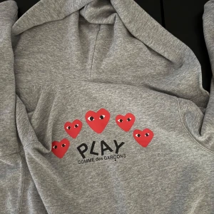 Grå hoodie Comme des Garçons Play - Grå hoodie från Comme des Garçons Play med dragkedja och huva. Framsidan har ett rött hjärta med ögon broderat på bröstet och baksidan är dekorerad med flera röda hjärtan och loggan. Mjuk bomullskänsla och klassisk passform.