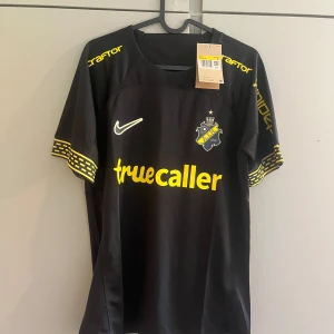 AIK svart matchtröja Nike 2024 - AIK:s officiella svarta matchtröja från Nike med gula detaljer och klubbmärke på bröstet. Truecaller och SVEA-loggor i gult, samt mönstrade ärmslut. Tillverkad i lätt och ventilerande polyester, perfekt för fotbollsplanen eller läktaren.