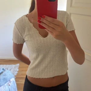 Guldig stickad croppad topp - Säljer en guldig stickad topp med korta ärmar och v-ringad hals. Toppen har ett vågigt avslut och ett snyggt strukturerat mönster i tyget. Perfekt till jeans eller kjol för en chill och trendig look.