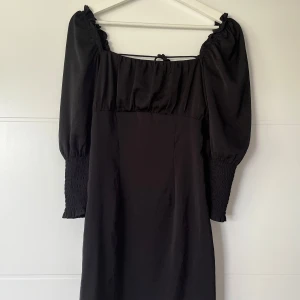 Svart klänning med snörning i ryggen - Supersnygg svart klänning med puffiga långärmar och smock vid ärmsluten. Klänningen har fyrkantig ringning och cool snörning i ryggen som ger en unik look. Perfekt för dig som gillar trendiga detaljer och en stilren vibe.