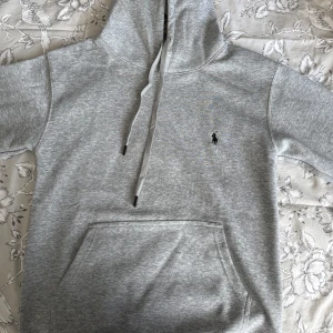 Grå hoodie från Ralph Lauren - Snygg grå hoodie från Ralph Lauren med klassisk broderad logga på bröstet. Tröjan har huva med snörning, känguruficka och är tillverkad i mjuk bomull. Perfekt för en chill och stilren look.