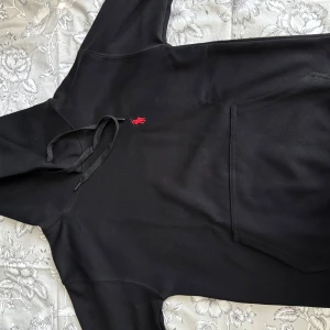 Svart hoodie från Polo Ralph Lauren - Svart hoodie från Polo Ralph Lauren med klassisk röd broderad logga på bröstet. Tröjan har huva med snörning och en stor magficka. Tillverkad i mjuk bomull, perfekt för en chill och stilren look.