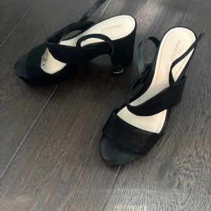 Svarta klackade sandaler från Mango - Stilrena svarta sandaler från Mango med breda korsade remmar och öppen tå. Skorna har en chunky klack och justerbar rem runt hälen. Tillverkade i mocka för en lyxig känsla och passar perfekt till sommarens outfits.