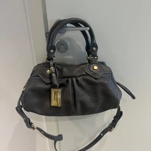 Marc Jacobs svart axelväska i skinn - Snygg svart axelväska från Marc Jacobs i mjukt skinn med guldfärgade metalldetaljer. Väskan har både handtag och justerbar axelrem samt dragkedja upptill. Insidan är fodrad med coolt svartvitt bokstavsmönster.