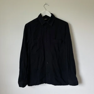Svart Arket overshirt i bomull, regular fit - Stilren svart overshirt i regular fit med klassisk krage och två stora bröstfickor. Skjortan är tillverkad i bomull och har långa ärmar med knappar vid manschetterna. Perfekt att bära öppen eller stängd för en avslappnad look. Artnr: C373