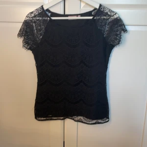 Svart spetsblus från KappAhl - Svart blus från KappAhl i storlek S med genombruten spets och korta ärmar. Blusen har ett snyggt vågformat spetsmönster och är lätt transparent på ärmarna. Perfekt för dig som gillar detaljer och en elegant look. jag skulle även säga att den passar Xs