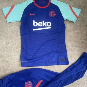 FC Barcelona Nike fotbollströja blå - FC Barcelona matchtröja från Nike i blått med ljusblå ärmar och röda detaljer. Tröjan har klubbmärke och Nike-logga på bröstet samt BEKO-tryck. Tillverkad i lätt funktionsmaterial, perfekt för träning eller match.