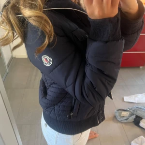 Mörkblå Moncler pufferjacka - Säljer en mörkblå pufferjacka från Moncler med klassisk logga på ärmen. Jackan har ribbade muddar, dragkedja och knappar framtill samt två bröstfickor och två sidofickor. Perfekt för kyliga dagar och riktigt snygg passform.