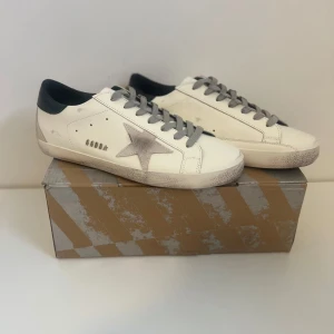 Golden Goose vita sneakers med stjärna - Snygga vita sneakers från Golden Goose med grå stjärndetalj på sidan, grå snörning och svart häl. Skorna har en vintage-inspirerad look med slitna detaljer och platt sula. Perfekta för dig som gillar streetstyle och unika detaljer.