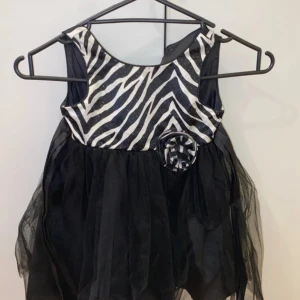 Zebramönstrad svart kortklänning - Supersöt kortklänning med zebramönstrad överdel i svart och vitt, dekorerad med en matchande blomdetalj i midjan. Kjolen är fluffig och gjord av svart tyll som ger extra volym. Perfekt för dig som vill sticka ut med en cool look.