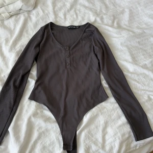 Brun ribbad body från PrettyLittleThing - Säljer en brun ribbad body från PrettyLittleThing med lång ärm och v-ringning. Toppen har tre knappar framtill och är figurnära med en snygg, croppad skärning vid höfterna. Perfekt att styla med jeans eller kjol för en clean look.