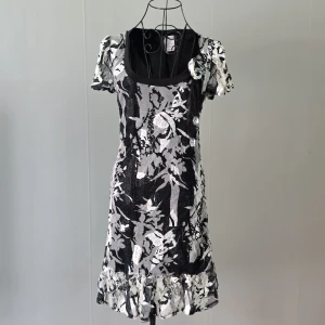 Svartvit blommig kortklänning - Unik svartvit kortklänning med blommigt mönster och volangkant nertill. Klänningen har korta ärmar, rundad urringning och en dekorativ blomdetalj vid axeln. Materialet har en lätt glittrig känsla som ger extra edge till looken.