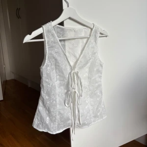Vit broderad topp med snörning - Superfin vit väst i bomull med broderade blommor och blad. Toppen har v-ringning och snygg snörning.