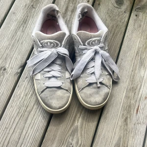 Grå Adidas Campus sneakers i mocka - Snygga Adidas Campus sneakers i ljusgrå mocka med klassiska vita ränder på sidorna och breda vita skosnören. Skorna har en platt sula och rund tå, samt rosa innersula för en extra touch. Trasiga bak så säljer de för lågt pris💓är även för stora för mig 