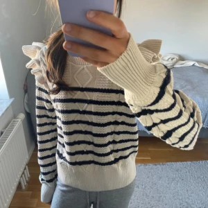 Randig stickad tröja Holly & Whyte XS - Supersöt stickad tröja från Holly & Whyte i krämvitt med mörkblå ränder. Tröjan har volangdetaljer på axlarna, kabelstickat mönster och ribbade muddar. Perfekt för dig som gillar en trendig och unik look med extra detaljer.