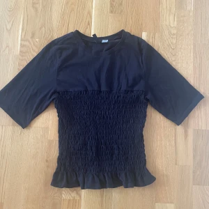 Svart smockad topp med volangkant - Svart topp med smockad midja och volangkant nertill. Sitter mycket fint på kroppen