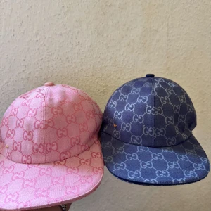 Gucci kepsar i rosa och blått - Två snygga Gucci kepsar med GG-monogram över hela kepsen. En i ljusrosa och en i marinblå, båda med böjd skärm och klassisk passform. Tillverkade i bomullscanvas med tydlig logga och justerbar baksida. Perfekt för dig som vill sticka ut med lyxig streetstyle.