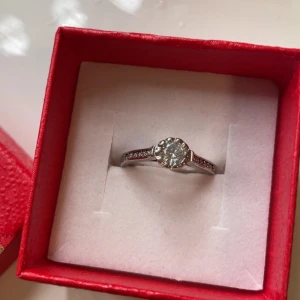 Silverring med glittrande sten - Säljer en snygg rostfritt silverfärgad ring med en stor rund glittrande sten i mitten och små stenar längs sidorna på ringen. Kommer i en röd ask. Inget fel på ringen.