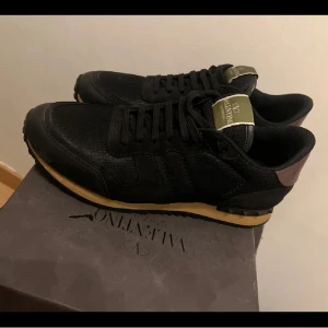 Valentino Garavani Rockrunner sneakers - Svarta Valentino Garavani Rockrunner sneakers med snygga detaljer i skinn och textil. Skorna har en beige sula, bruna hälkappor och ikoniska nitar baktill. Klassisk snörning och logga på plösen. Perfekta för dig som vill ha en lyxig och stilren sneaker. Box två dustbags valentino kort och extra skosnöre kommer med 