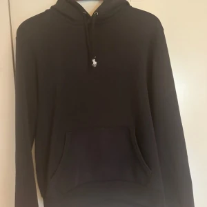 Mörk blå hoodie från Ralph Lauren - Mörk blå hoodie från Ralph Lauren med klassisk vit broderad logga på bröstet. Tröjan har huva med snörning och en  magficka framtill. Tillverkad i mjuk bomull och har lång ärm. Perfekt för en chill och stilren look.