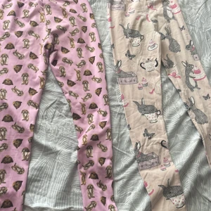 2 par leggings med kaninmotiv - Två par mjuka leggings från Mywear. Ena paret är ljusrosa med gulligt kaninmönster i brunt och beige, det andra paret är ljusbeige med tecknade kaniner, tekoppar och fjärilar i svart, rosa och grått. Båda har resår i midjan och är perfekta för chill dagar.