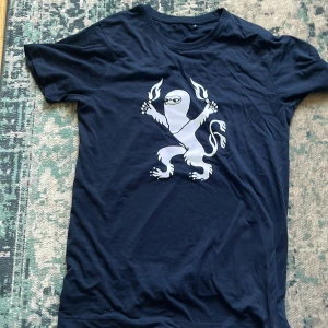 Mörkblå t-shirt från kommandobryggan Blåvitt  - Snygg mörkblå t-shirt i bomull med ett unikt vitt tryck av ett lejon  som håller två bangalrg Klassisk rund hals och korta ärmar, perfekt för dig som älskar blåvitt 