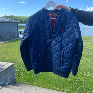 Blå bomberjacka från Polo Ralph Lauren - Snygg mörkblå quiltad bomberjacka från Polo Ralph Lauren med röd logga på bröstet. Jackan har dragkedja, ribbade muddar och två fickor med tryckknappar. Insidan är röd för en extra cool kontrast. Perfekt för höst och vår! Storlek 14-16 år. 
