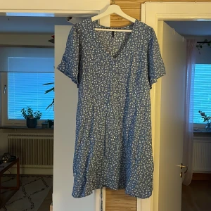 Blå blommig kortklänning från xlnt - Supersöt blå kortklänning från xlnt med vitt blommönster. Klänningen har v-ringning, korta ärmar och knytband i midjan för en snygg passform. Perfekt för varma dagar och har en luftig känsla tack vare det tunna bomullstyget.