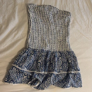 Vit och blå kortklänning Gina Tricot - Supersöt axelbandslös kortklänning från Gina Tricot i vitt och blått. Smockad överdel och volangig kjol med unikt mönster. Perfekt för varma dagar och sommarhäng. Klänningen har fina spetsdetaljer längs volangerna och är lätt och luftig.