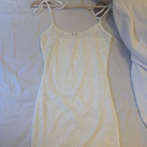 Vit spetsklänning med smala axelband - Supersöt vit kortklänning med smala axelband som knyts på axlarna. Klänningen har ett vackert spetsmönster över hela och är figurnära med en feminin känsla. Perfekt för sommarens alla tillfällen!