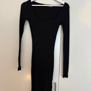 Svart ribbad klänning från NA-KD - Svart ribbad kortklänning från NA-KD med lång ärm och fyrkantig ringning. Klänningen är tight och har en stilren, minimalistisk look som passar perfekt till sneakers eller boots. Materialet är mjukt och stretchigt för en bekväm passform.