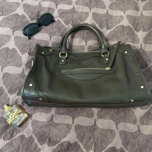 Svart handväska i skinn med detaljer - Snygg svart handväska i skinn med guldfärgade metalldetaljer och dragkedja framtill. Väskan har två rejäla handtag och rymligt innerfack med flera fack för småsaker. Perfekt för dig som vill ha en stilren och praktisk väska med coola detaljer.